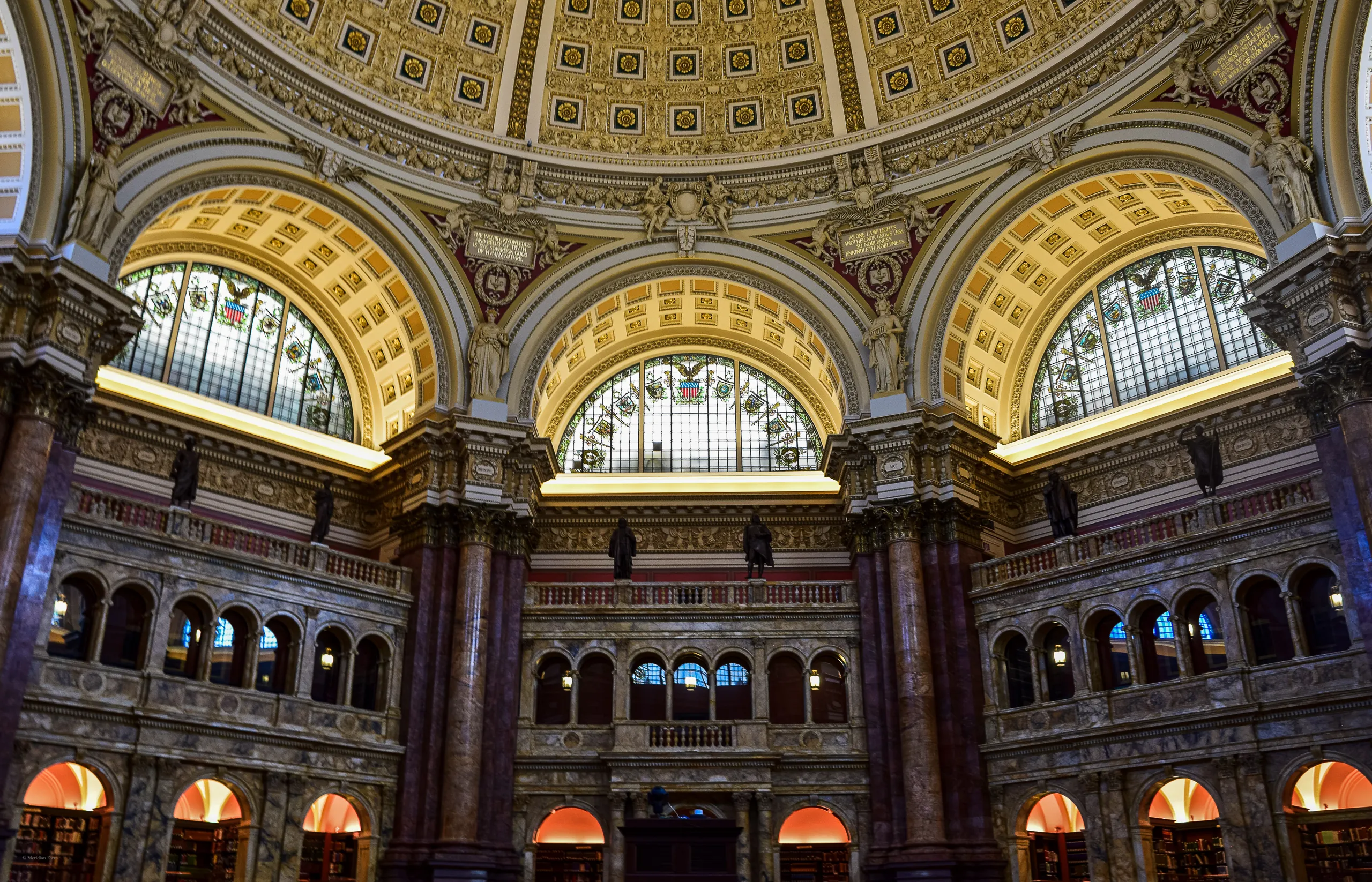 The Main Reading Room of the Library of Congress: three arched stained-glass windows beneath a gilded coffered dome, framed by marble columns and tiers of arches below.